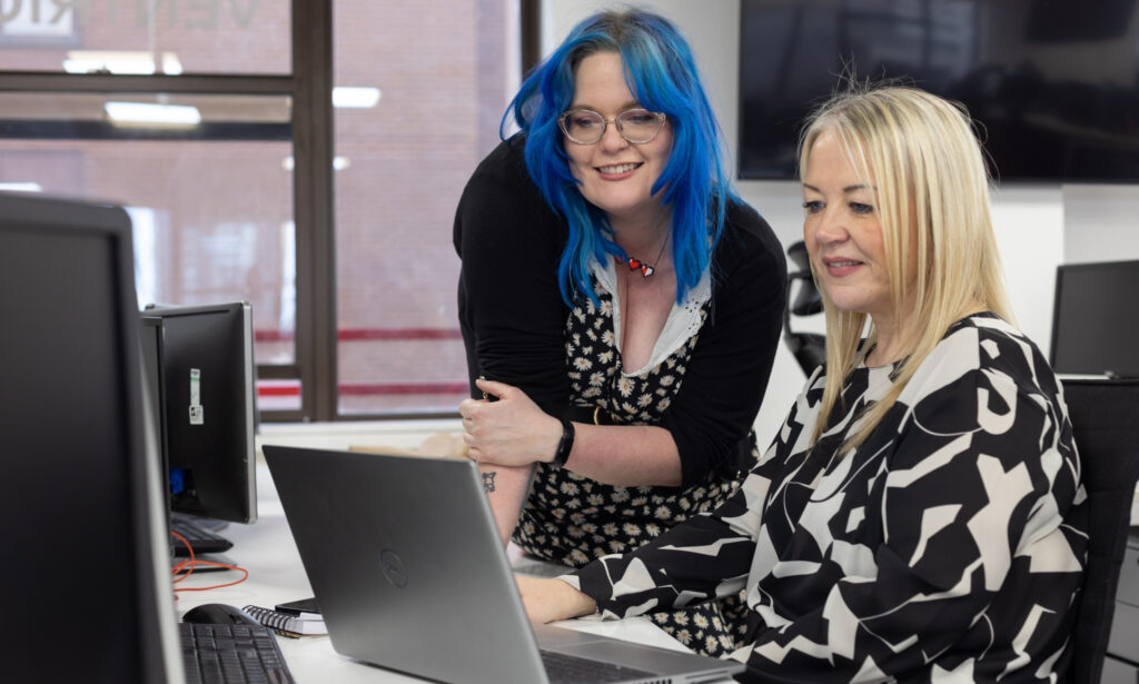 Two female colleagues consulting at a desk (one seated, one leaning)